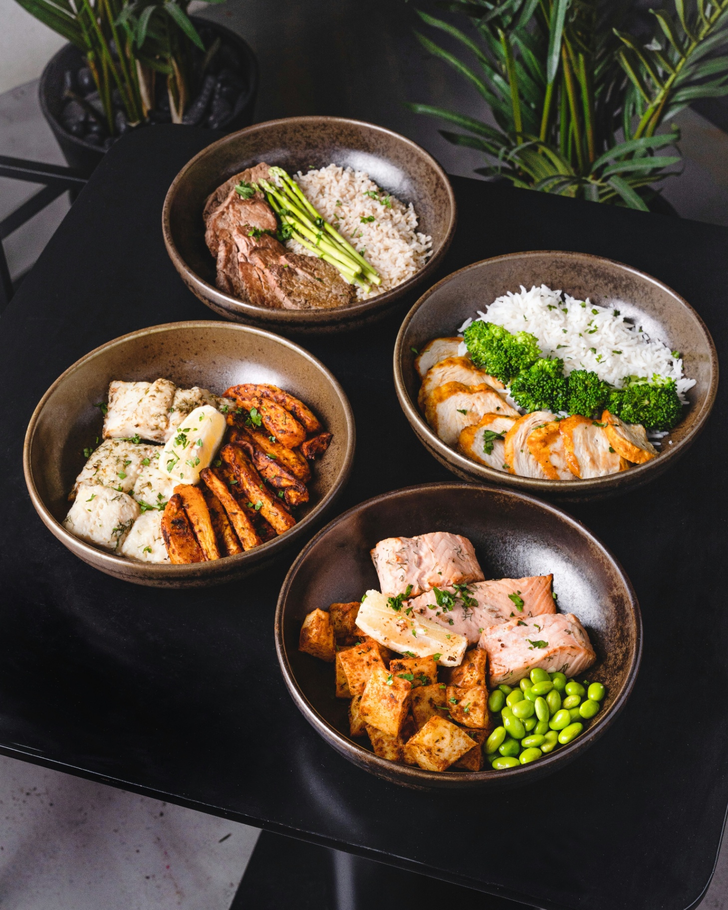 Four seasonal grain bowls with salmon, chicken, steak, and fish, plated on dark ceramic.